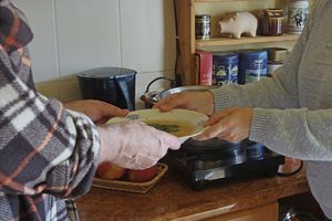 Na zdjęciu dwie ręce wymieniają talerz z zupą, w tle kuchnia z akcesoriami.
