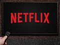 Obrazek przedstawia ekran telewizora z logo Netflix na czarnym tle, ręka trzymająca pilot.