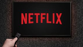 Obrazek przedstawia ekran telewizora z logo Netflix na czarnym tle, ręka trzymająca pilot.