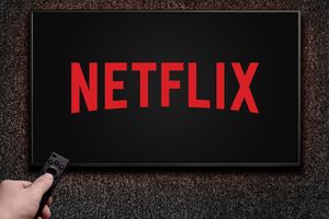 Obrazek przedstawia ekran telewizora z logo Netflix na czarnym tle, ręka trzymająca pilot.