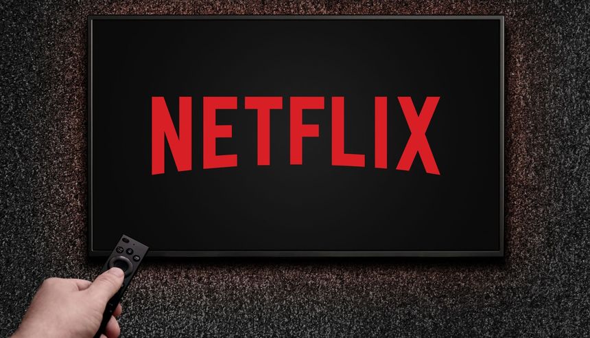 Obrazek przedstawia ekran telewizora z logo Netflix na czarnym tle, ręka trzymająca pilot.