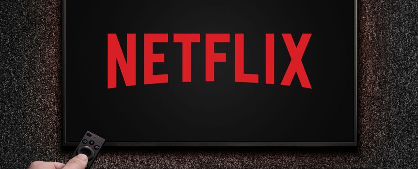 Obrazek przedstawia ekran telewizora z logo Netflix na czarnym tle, ręka trzymająca pilot.