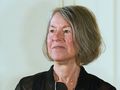 Louise Glück. Kobieta z krótkimi, siwymi włosami, uśmiechnięta, w czarnej bluzie, stoi na tle jasnej ściany.