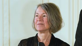 Louise Glück. Kobieta z krótkimi, siwymi włosami, uśmiechnięta, w czarnej bluzie, stoi na tle jasnej ściany.