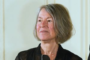 Louise Glück. Kobieta z krótkimi, siwymi włosami, uśmiechnięta, w czarnej bluzie, stoi na tle jasnej ściany.