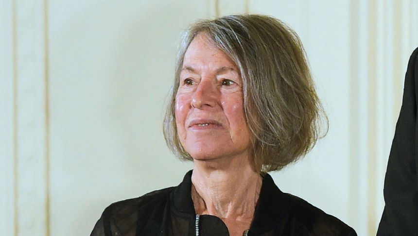 Louise Glück. Kobieta z krótkimi, siwymi włosami, uśmiechnięta, w czarnej bluzie, stoi na tle jasnej ściany.