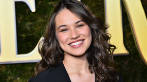 Młoda kobieta z kręconymi włosami uśmiecha się, stoi na tle zielonej ściany z napisami.