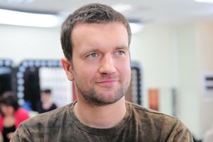 Mężczyzna z krótkimi, ciemnymi włosami i zielonymi oczami, ubrany w brązowy t-shirt.