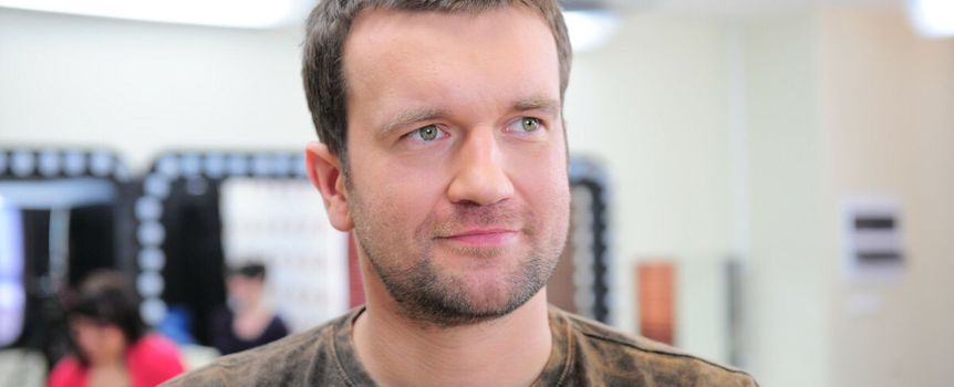 Mężczyzna z krótkimi, ciemnymi włosami i zielonymi oczami, ubrany w brązowy t-shirt.
