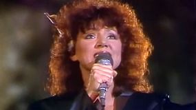 Kobieta z kręconymi włosami trzyma mikrofon, śpiewa na scenie z emocjonalnym wyrazem twarzy.