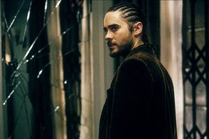 Mężczyzna z warkoczami patrzy przez pęknięte lustro, w tle widać ciemne wnętrze.