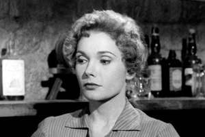 Kobieta z krótkimi, falowanymi włosami w pasiastym ubraniu, w tle półki z butelkami.