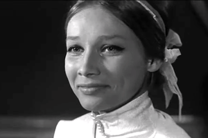 Młoda kobieta w białej bluzie, uśmiechająca się, z włosami spiętymi w kok. Tło ciemne.