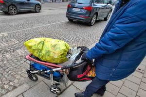 Mężczyzna w niebieskiej kurtce prowadzi wózek z żółtym workiem pełnym rzeczy. Ulica z kostki brukowej.