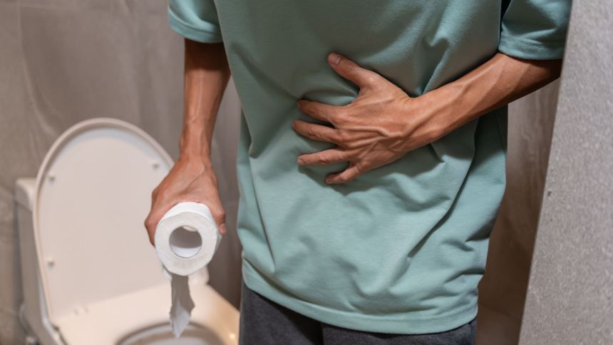 Mężczyzna w t-shircie trzyma papier toaletowy, trzymając się za brzuch przy toalecie.