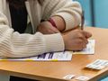 Na zdjęciu widoczna jest ręka osoby w białym swetrze, która pisze na kartce przy biurku.