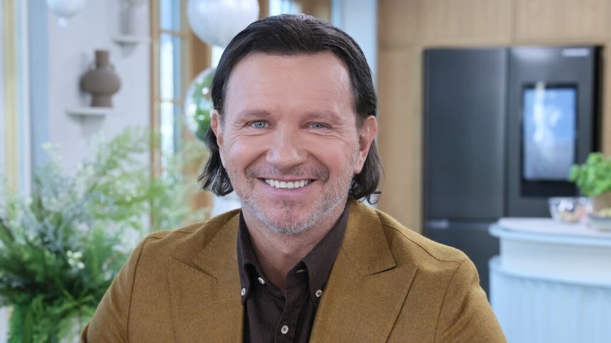 Uśmiechnięty Radosław Majdan w brązowym garniturze, siedzący w jasnym pomieszczeniu.