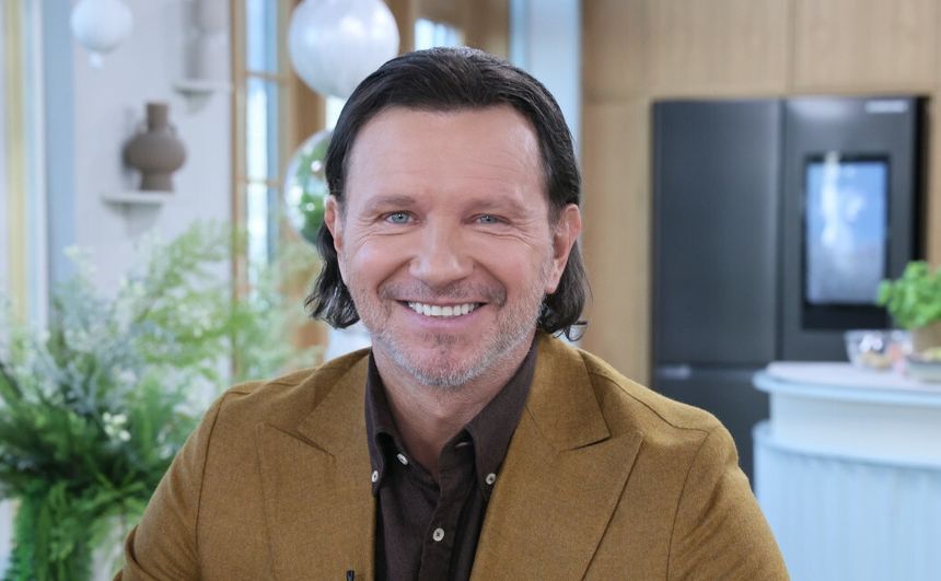 Uśmiechnięty Radosław Majdan w brązowym garniturze, siedzący w jasnym pomieszczeniu.