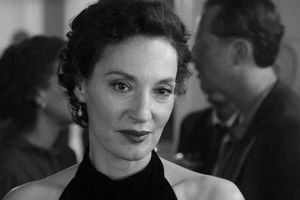 Kobieta o krótkich, ciemnych włosach w czarnej sukience, uśmiecha się w eleganckim otoczeniu.