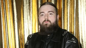 Mężczyzna z długą brodą w czarnej skórzanej kurtce, na tle złotych zasłon.