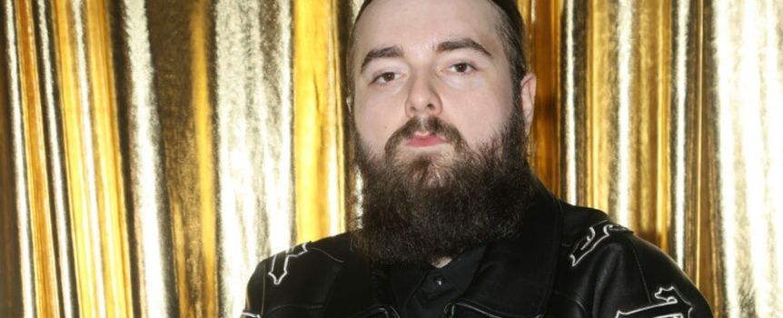 Mężczyzna z długą brodą w czarnej skórzanej kurtce, na tle złotych zasłon.