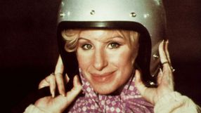 Kobieta w hełmie motocyklowym uśmiecha się, trzymając go w dłoniach, z kolorowym szalikiem.