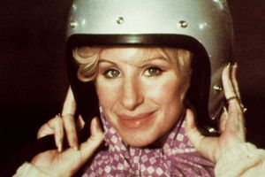 Kobieta w hełmie motocyklowym uśmiecha się, trzymając go w dłoniach, z kolorowym szalikiem.