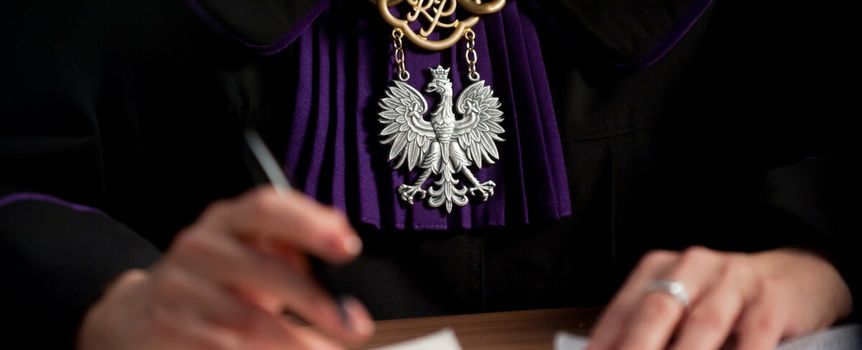 Sędzia w togach, piszący dokumenty, z godłem Polski widocznym na medalu.