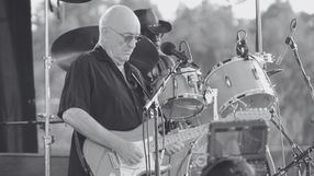 Muzyk grający na elektrycznej gitarze na tle perkusji, w czarno-białej tonacji.