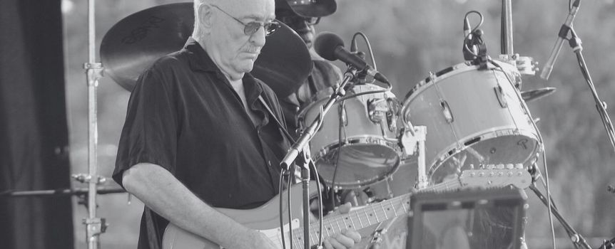 Muzyk grający na elektrycznej gitarze na tle perkusji, w czarno-białej tonacji.