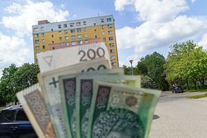Na pierwszym planie banknoty w dłoni, w tle blok mieszkalny oraz zielone drzewa.