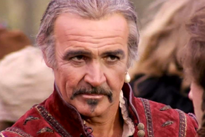 Mężczyzna w czerwonym stroju z perłą na uchu, z intensywnym wyrazem twarzy. Tło rozmyte.