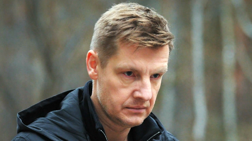 Mężczyzna w czarnej kurtce z poważnym wyrazem twarzy, w tle zieleń lasu.