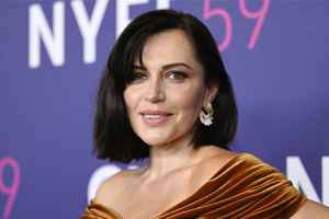 Kobieta w brązowej sukni z dekoltem, z krótkimi włosami i dużymi, ozdobnymi kolczykami.