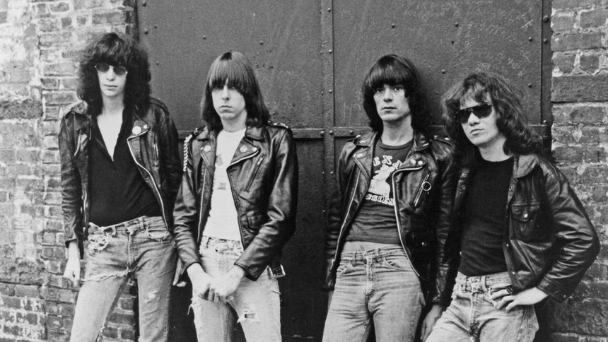 Czterech mężczyzn w skórzanych kurtkach, z okularami przeciwsłonecznymi, stojących na tle ceglanej ściany.