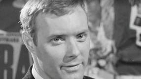 Mężczyzna w garniturze z krótkimi włosami, patrzy w obiektyw, w tle różne plakaty.