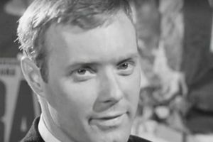 Mężczyzna w garniturze z krótkimi włosami, patrzy w obiektyw, w tle różne plakaty.