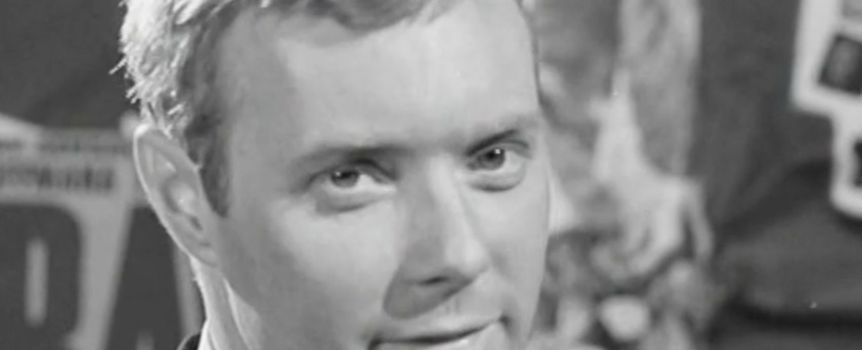 Mężczyzna w garniturze z krótkimi włosami, patrzy w obiektyw, w tle różne plakaty.