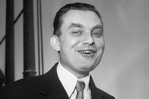 Mężczyzna w garniturze, uśmiechnięty, z krótkimi włosami, stoi na tle neutralnego tła.