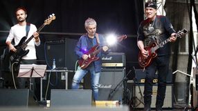 Zespół muzyczny grający na scenie, trzech muzyków z gitarami i basem, otoczonych sprzętem.