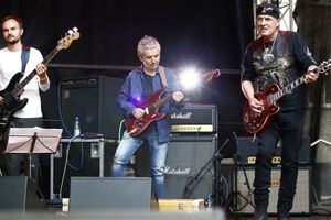 Zespół muzyczny grający na scenie, trzech muzyków z gitarami i basem, otoczonych sprzętem.