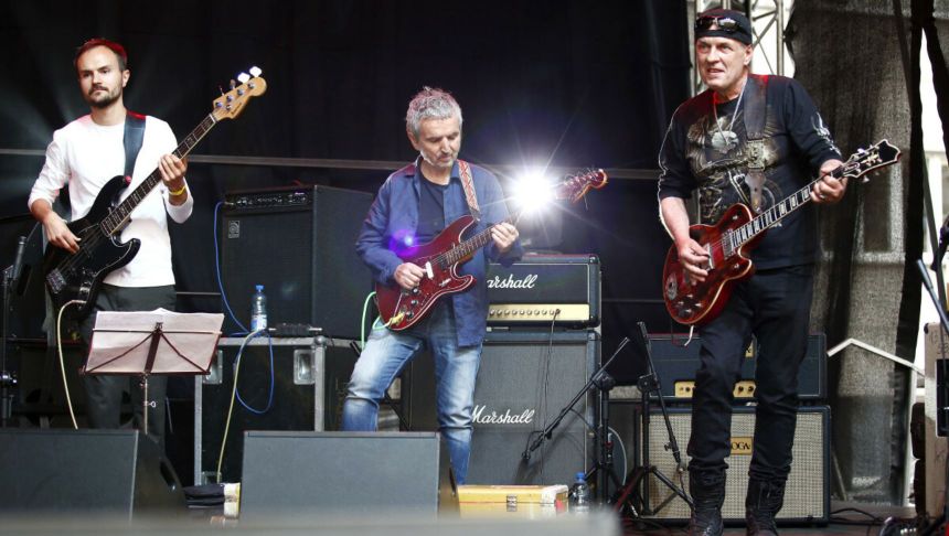 Zespół muzyczny grający na scenie, trzech muzyków z gitarami i basem, otoczonych sprzętem.