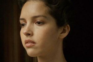 Kobieta o krótkich włosach, zamyślonym wyrazie twarzy, w tle ciemne zasłony.