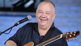 Mężczyzna z uśmiechem trzyma gitarę, występując w studiu z niebieskim tłem.