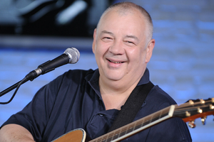 Mężczyzna z uśmiechem trzyma gitarę, występując w studiu z niebieskim tłem.