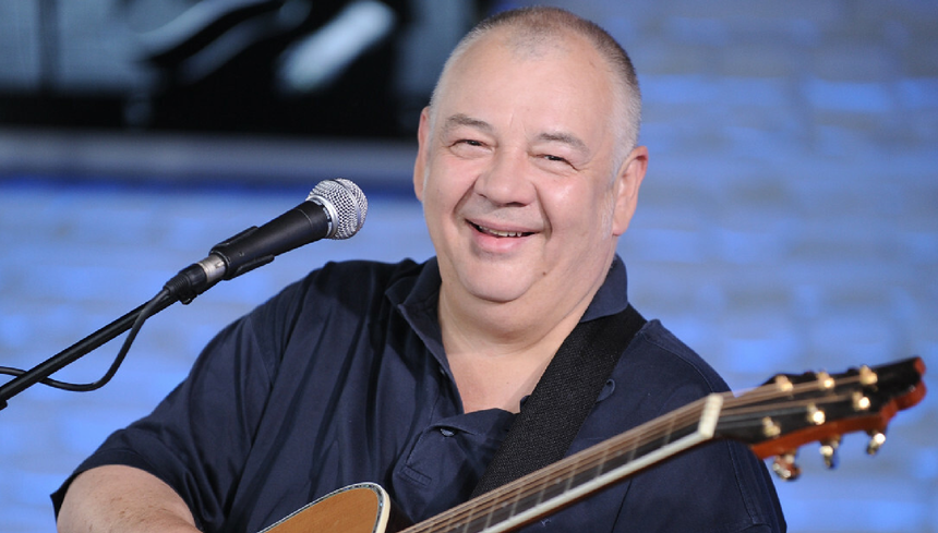 Mężczyzna z uśmiechem trzyma gitarę, występując w studiu z niebieskim tłem.