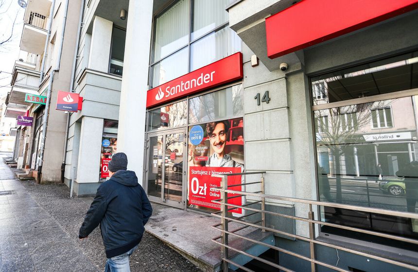 Osoba w ciemnej kurtce idzie w kierunku banku Santander z reklamą na szkle.