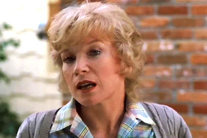 Kobieta z krótkimi, kręconymi włosami, w koszuli w kratę, z tłem z ceglanych ścian.