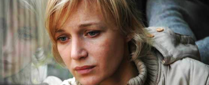 Kobieta o blond włosach w beżowej kurtce wygląda smutno, patrząc w dal przez szkło.