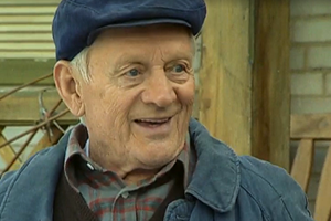 Na zdjęciu uśmiechnięty starszy mężczyzna w granatowej czapce i kurtce. Tło drewniane.